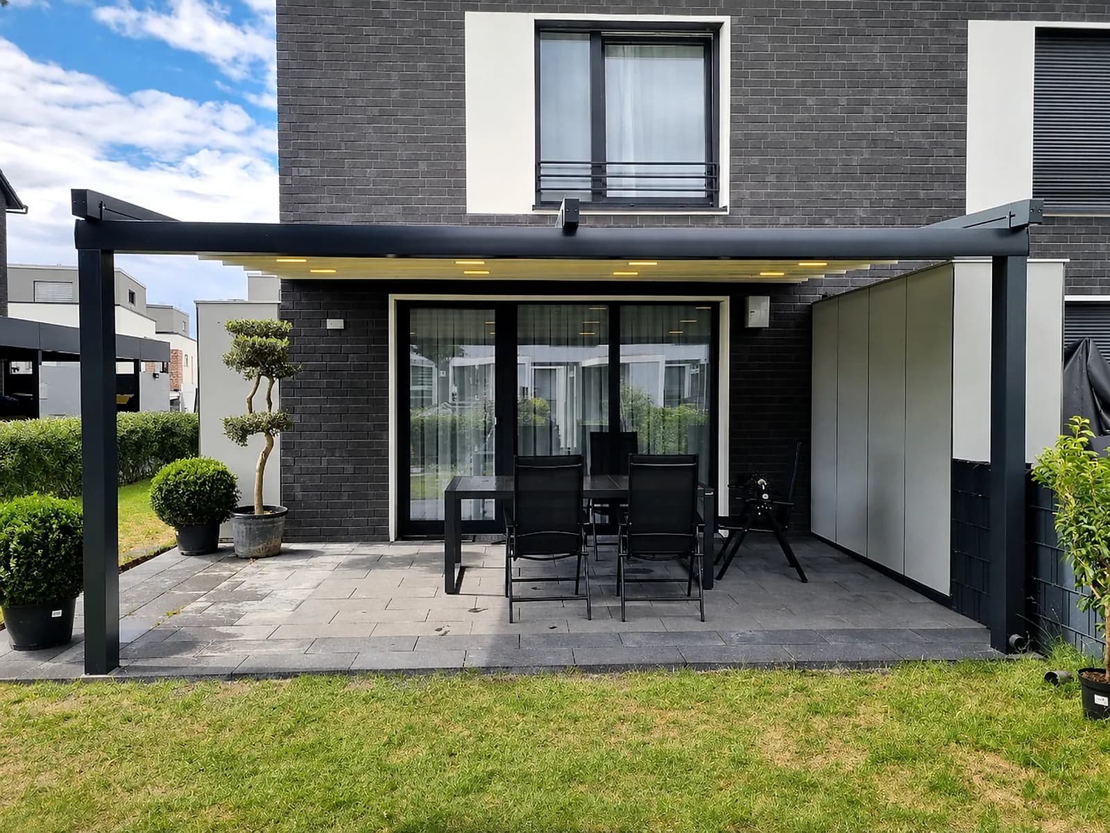 Pergola mit Beleuchtung über der Terrasse eines dunklen Klinkerhauses